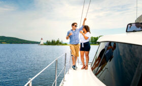 Couple enjoys a drink on the deck of the Cape Bretoner 1 catamaran with Baddeck Bay Lighthouse in the background